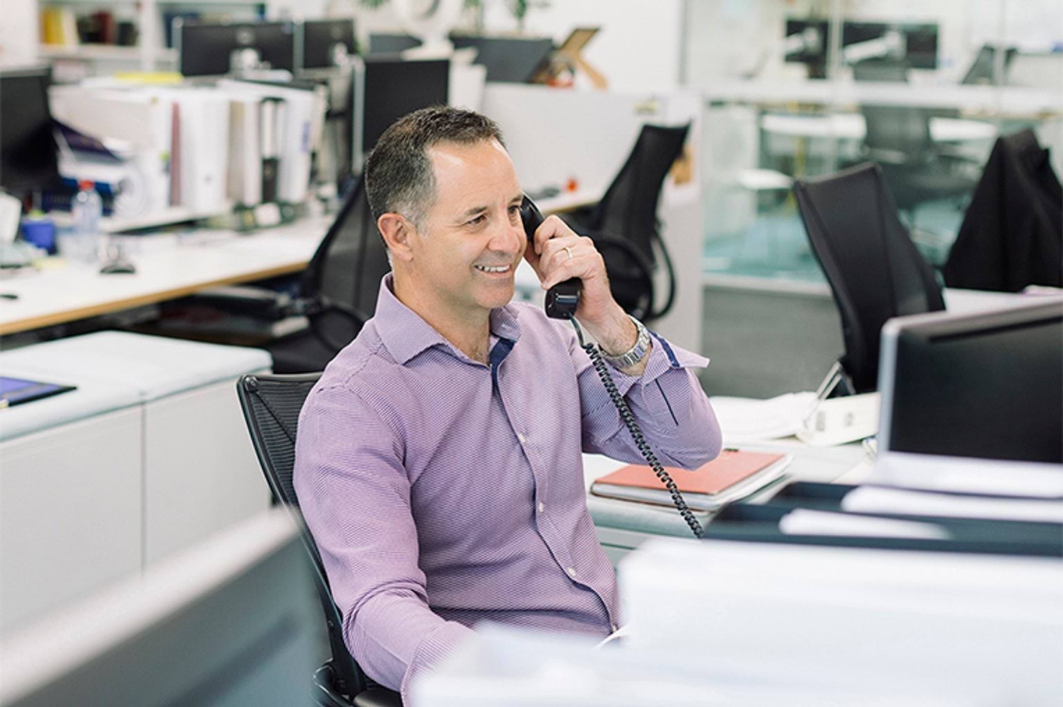 A photograph of a person talking on the phone at a desk in an office. 