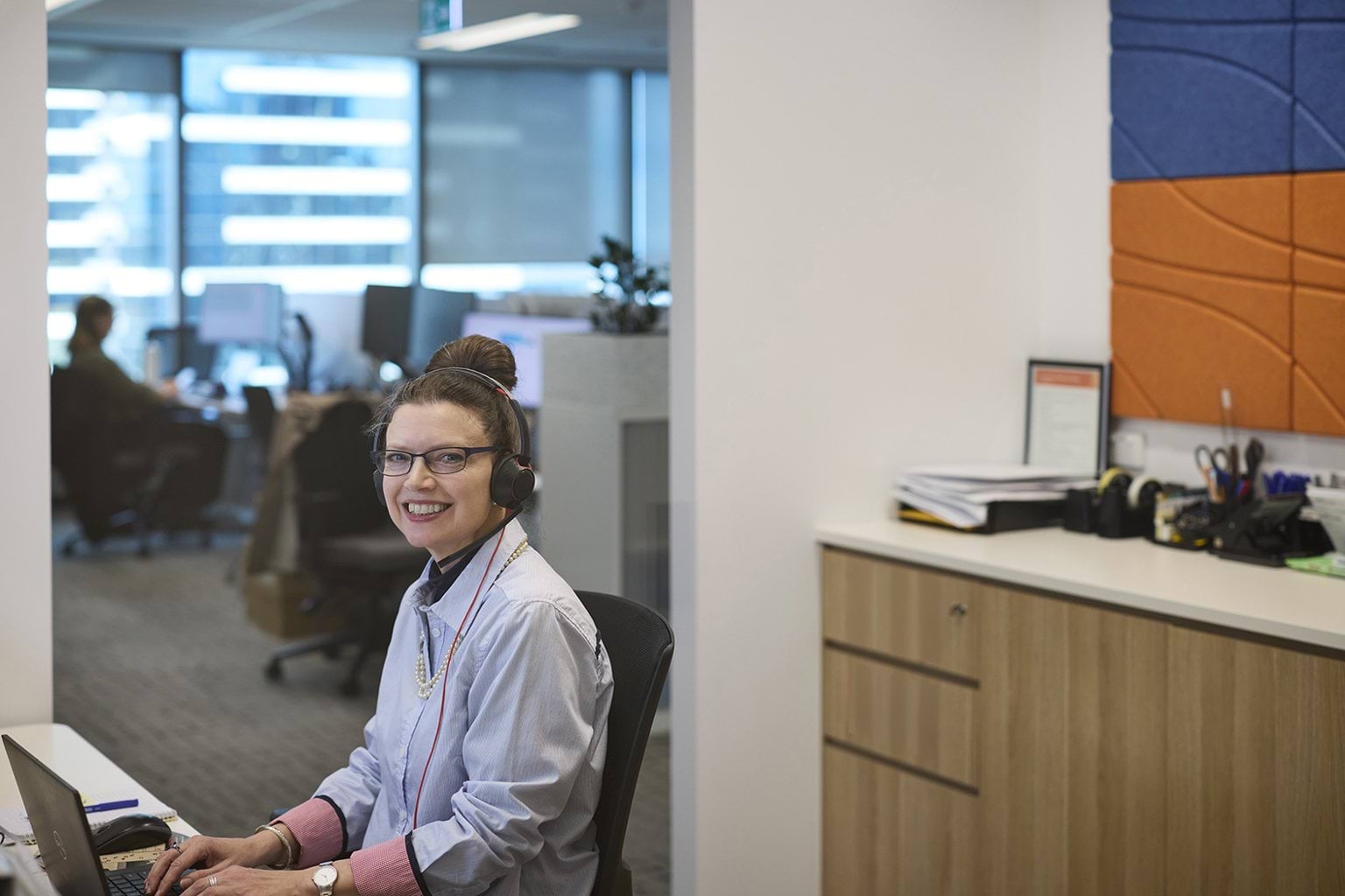 A person at a desk in an office wearing a headset and smiling. 